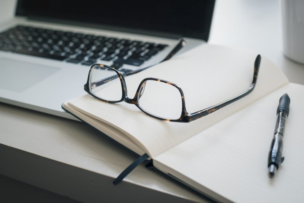 a desk with laptop, glasses and a notebook