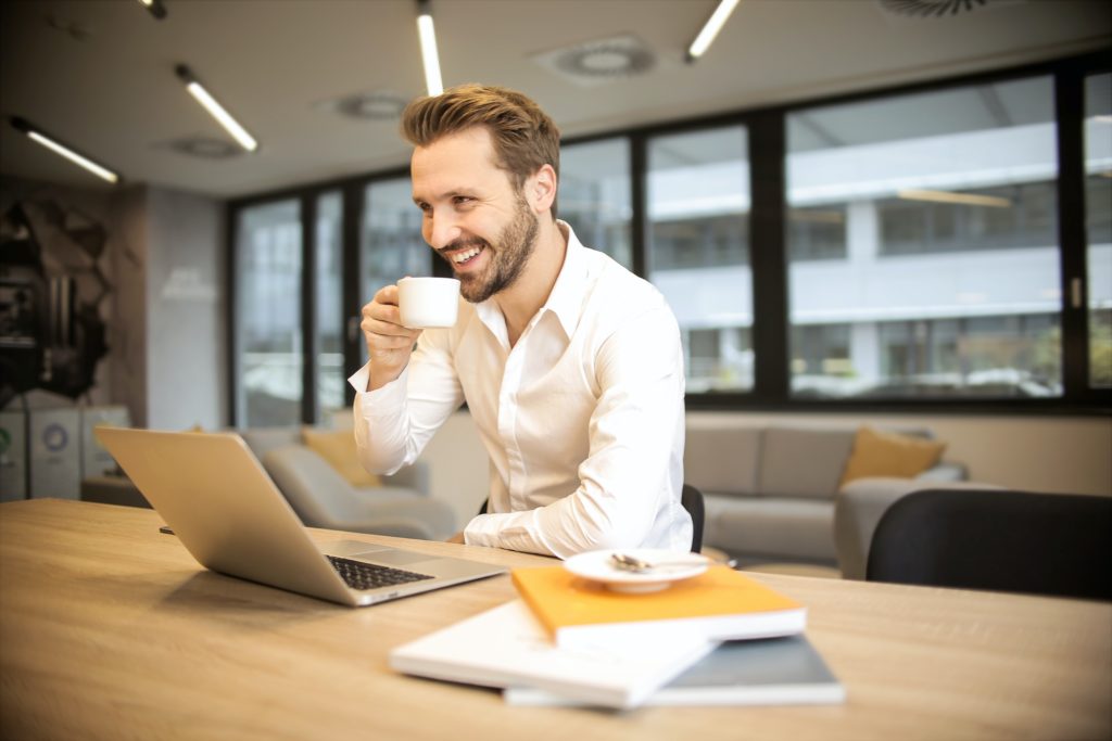 man stilling at desk drinking coffee and smiling at laptop
